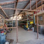 A storage shed with metal fencing, shelves holding tools, bins, and equipment, and various items organized along the sides. Gardening tools and barrels are visible on the left, and large shelves extend to the back.