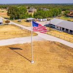 An American flag on a tall flagpole stands in the center of a dry, grassy area with a paved driveway, surrounded by houses, fences, and trees in a rural neighborhood.