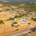 Aerial view of a rural residential area with scattered houses, barns, a pond, winding driveways, and dry grassy fields bordered by a road and surrounded by trees and hills in the background.