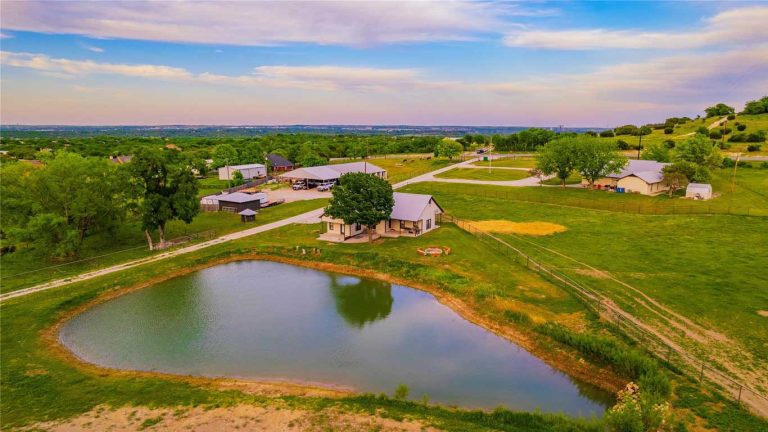 A small farmhouse with a gray roof sits by a pond, surrounded by green fields, trees, and farm buildings under a colorful sky, with a scenic rural landscape in the background.