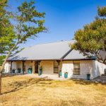 A single-story house with a metal roof and white stone exterior sits on a dry, grassy yard with several trees providing shade. The home features a covered front porch with green potted plants and clear blue sky above.