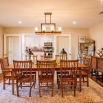 A warm, inviting dining room with a wooden table and eight matching chairs, a rug beneath, a sideboard, wall art reading “home,” and a view into a bright kitchen in the background.