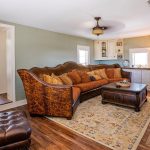 A cozy living room with a brown sectional sofa, patterned rug, wooden coffee table, cushioned chair, TV mounted above a fireplace, and a windowed corner cabinet with a lamp. A bedroom is visible through an open doorway.