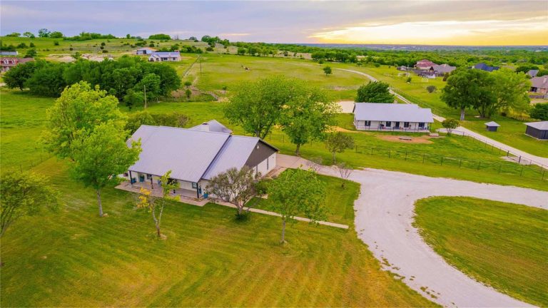 Aerial view of a ranch with a main house, a barn, trees, and a curved gravel driveway, surrounded by green fields and neighboring properties under a partly cloudy sunset sky.