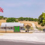 A metal gate with a large green "M" stands at the entrance to a ranch. An American flag waves on the left, and a gravel driveway leads to a farmhouse and trees under a clear sky.