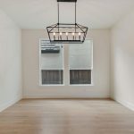 A bright, empty room with light wood floors, white walls, two large windows, and a modern black rectangular chandelier hanging from the ceiling.