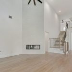 Spacious modern living area with light wood floors, tall white walls, a high ceiling with a black ceiling fan, a double-sided fireplace, and an open staircase leading to an upper level.
