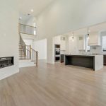 Spacious modern open-concept living area with light wood floors, a built-in wall fireplace, a large kitchen with island, and a staircase leading to an upper level. The room features high vaulted ceilings and ample natural light.