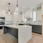 Modern kitchen with a large marble island, pendant lights, built-in ovens, white cabinets, dark lower cabinets, and an empty space for a refrigerator. Light wood floors and bright natural lighting fill the room.