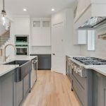Modern kitchen with white and gray cabinets, stainless steel appliances, a farmhouse sink, a gas stove, marble countertops, light wood flooring, and a staircase visible in the background.