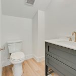 A modern bathroom with white walls, a white toilet, a dark gray vanity with a white countertop, gold faucet fixtures, and light wood flooring.