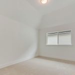 A small, empty room with light-colored walls and carpet, a single window letting in natural light, and a ceiling light fixture. The room has a sloped ceiling and a clean, minimal appearance.