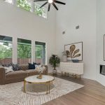 A modern living room with high ceilings, large windows, neutral sofas, a round coffee table, light wood floors, and wall art above a minimalist fireplace. Greenery and soft natural light fill the space.