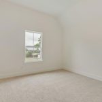 An empty, unfurnished room with white walls, beige carpet flooring, and a single window letting in natural light. The window provides a view of greenery and a cloudy sky outside.