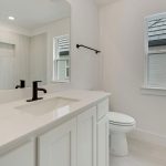 A modern bathroom with a white vanity, black faucet, large mirror, toilet, and window. The space has light-colored walls and floor tiles, creating a clean and minimalistic look.