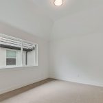 A bright, empty room with white walls, beige carpet, a single ceiling light, and a large window showing the exterior of a neighboring building.