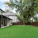 A modern two-story house with large windows and a covered patio overlooks a green backyard with a tall tree and a wooden privacy fence. The sky is partly cloudy.