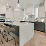 Modern kitchen with a large marble island, three woven barstools, stainless steel appliances, white upper cabinets, dark lower cabinets, pendant lights, and light wood flooring. A hallway and door are visible in the background.