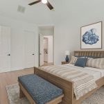 A modern bedroom with a wooden bed, blue and beige bedding, a matching bench, two nightstands with blue lamps, a dresser with decor, and wall art of a blue flower above the bed. The room has light walls and hardwood floors.
