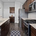 Modern kitchen with dark wood cabinets, stainless steel appliances, granite countertops, a double sink, gas stove, and white tile backsplash. A small geometric rug lies on the dark wood floor.