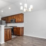 A modern kitchen with wood cabinets, stainless steel appliances, pendant lighting over a counter, and an adjacent dining area with a chandelier. The floor is dark wood, and the walls are light-colored.
