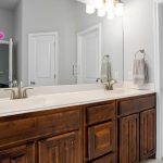 A modern bathroom features a double sink with wooden cabinets, a large mirror, three overhead lights, a white countertop with a potted orchid, and a towel hanging by a closed white door.