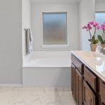 Modern bathroom with a white bathtub, double sink vanity with wooden cabinets, pink orchid on the counter, towel rack, and a glass shower with a brass handle; two frosted windows provide natural light.