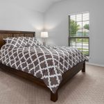 A neatly made bed with a brown and white patterned comforter sits in a bright bedroom with light walls, beige carpet, two bedside lamps, a window with blinds, and a flat-screen TV on a stand.