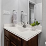 A modern bathroom features a dark wood vanity with a white countertop, a stainless steel faucet, a small potted plant, two hand towels on a ring, a large wall mirror, and light gray walls.