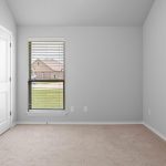 A small, empty room with light beige carpet, light gray walls, two white doors, and a window with blinds looking out to a grassy yard and houses.