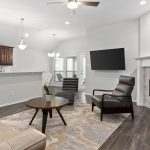 Modern living room with a tan sofa, armchair, round coffee table, wall-mounted TV, fireplace, and open kitchen with dark cabinets. Wooden flooring and pendant lights complete the bright, neutral space.