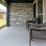 Two wicker chairs sit on a covered concrete patio outside a brick house, with a grassy yard and wooden fence visible in the background.