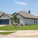 A single-story suburban house with gray brick exterior, dark trim, a two-car garage, and a neatly landscaped front yard with small trees and green grass, located on a corner lot.