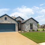 Single-story brick house with a dark blue garage door, well-kept lawn, and a landscaped garden bed, set in a suburban neighborhood under a partly cloudy sky.