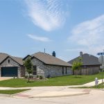 A suburban neighborhood features single-story brick houses with neatly trimmed lawns on a sunny day. A street sign and fire hydrant are visible at the intersection in the foreground.