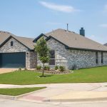 A single-story brick house with a dark garage door, neatly trimmed lawn, small trees, and landscaped flowerbeds on a sunny day in a suburban neighborhood.