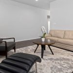 Modern living room with a beige sofa, black armchair, matching ottoman, round coffee table with a potted orchid, light patterned rug, and fireplace with a lit fire on the left, all against white walls.