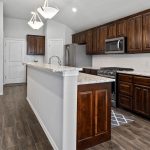 Modern kitchen with dark wood cabinets, granite countertops, stainless steel appliances, and a dual pendant light fixture. The space features a tile floor and an adjacent beige sofa in an open floor plan.