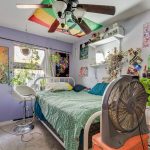 A colorful, cluttered bedroom with plants, posters, and art on lavender walls. A bed with a green quilt sits under a ceiling fan, and a large fan is on a dresser beside the bed. Sunlight streams through a window.