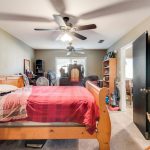 A cozy bedroom with a wooden bed, red plaid bedding, dresser, open closet door, and ceiling fans. Sunlight enters through a window with white blinds at the far end, and various furniture is arranged around the room.