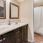 A bathroom with a double sink vanity, two mirrors above each sink, a toothbrush holder, and a soap dispenser. In the background, there is a shower with a white curtain and tiled flooring.