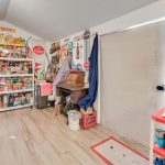 A cluttered room with shelves full of food, boxes, and vintage decor. An old wooden desk and chair sit against the wall, and a plastic cart with art supplies stands nearby. Light wood flooring and a closed door are visible.