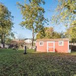 A backyard with green grass, tall trees, and a red shed with white trim. Shadows from the trees cover parts of the yard, and a wooden fence runs along the back and right side.