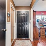 A view of a home's entryway with a black front door, brown and white walls, wooden floors, and a patterned rug. To the right, an office area with a wooden desk, chair, and plants is visible through an open doorway.