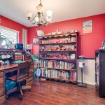A cozy home office with red walls, wooden desk and chair, bookshelf filled with books and collectibles, potted plants on the windowsill, and a chandelier hanging from the ceiling. Natural light streams in from the window.