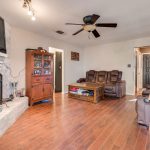 A cozy living room with wood flooring, brown leather recliners, a wooden coffee table, shelves, a brick fireplace with a TV mounted above, and a ceiling fan with lights. The front door is visible in the background.