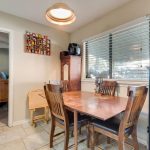 A cozy dining area with a wooden table and four chairs, tile flooring, a window with blinds, various cabinets, wall decorations, and a light hanging above the table. A doorway leads to another room with wooden furniture.