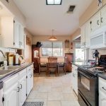 Bright kitchen with white cabinets, black countertops, double sink, stove, microwave, and neatly arranged items. In the background, there’s a dining table with chairs next to a window and a wooden hutch.