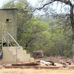 A small, weathered green hunting blind stands on stilts with concrete steps and a metal railing leading to its door. It is surrounded by trees and scattered debris in a grassy, rural area.