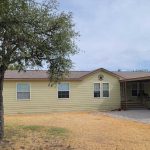A single-story beige house with brown trim, a covered porch, and a metal star decoration on the front. A white truck is parked under a carport, and there are trees and a patchy yellow lawn in the yard.